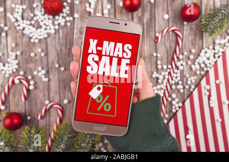 Concetto per Natale stagionale shopping online e vendita con mano azienda telefono cellulare con rosso "X-mas vendita " segno di Fronte desk con decora stagionali Foto Stock
