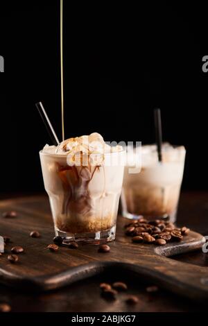 Liquore al caffè versando in white russian cocktail in bicchieri con cannucce su tavola di legno con i chicchi di caffè isolato su nero Foto Stock