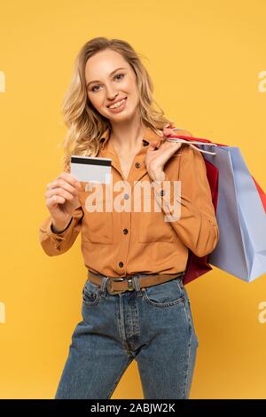 Donna sorridente che mostra la carta di credito e azienda shopping bags isolato su giallo Foto Stock