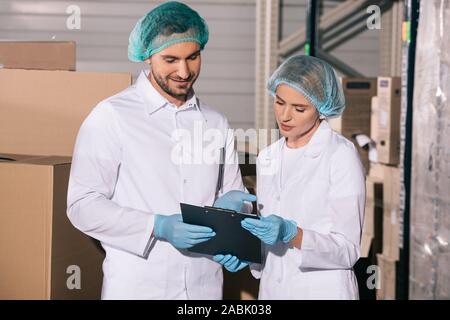 Due positive ammassatori in camici e retine per capelli guardando negli appunti in magazzino Foto Stock