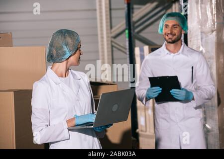 Sorridente ammassatore holding negli appunti e guardando il collega utilizzando laptop in magazzino Foto Stock