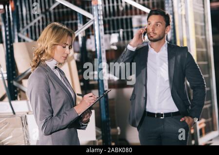 Imprenditrice concentrato sulla scrittura di appunti nei pressi di imprenditore parlando sullo smartphone in magazzino Foto Stock