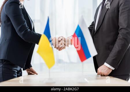 Vista ritagliata di diplomatici in abbigliamento formale stringono le mani nei pressi di ucraini e russi bandiere Foto Stock