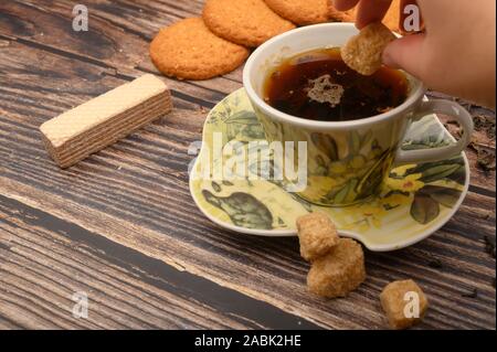 La mano della ragazza mette un pezzo di zucchero di canna in una tazza di tè nero, fiocchi d'avena cookies, foglie di tè, waffle, lo zucchero di canna su uno sfondo di legno. Close up Foto Stock