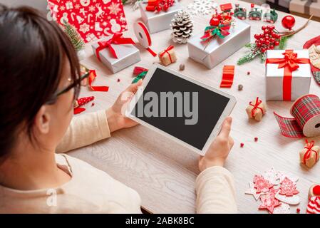 Ragazza con tablet circondato da decorazione di Natale e regali sul tavolo. Lo schermo è vuoto per il messaggio di saluto del testo o mockup, app o sito web design presentati Foto Stock