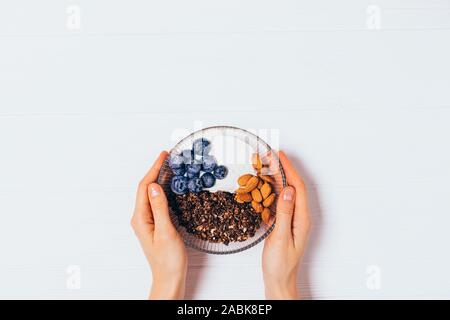 Mani femminili recipiente di contenimento del cioccolato artigianale di muesli con yogurt greco, mirtilli e mandorle bianche sul tavolo di legno, vista dall'alto. Foto Stock