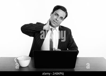 Studio shot del giovane imprenditore persiano seduta pur avendo dolore al collo con il computer portatile e la tazza di caffè sul tavolo in legno Foto Stock