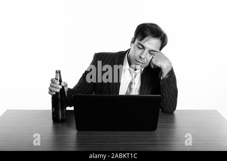 Studio shot del giovane imprenditore persiano cercando ubriaco mentre si tiene una bottiglia di birra e di seduta con il computer portatile sul tavolo di legno Foto Stock