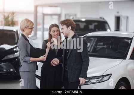 Di acquistare la loro prima vettura insieme. Angolo di Alta Vista del giovane venditore auto in piedi presso la concessionaria dicendo circa le caratteristiche della vettura per il Foto Stock