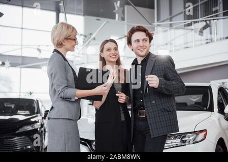 Di acquistare la loro prima vettura insieme. Angolo di Alta Vista del giovane venditore auto in piedi presso la concessionaria dicendo circa le caratteristiche della vettura per il Foto Stock