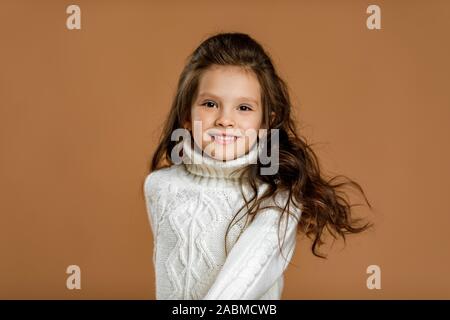 Carino sorridente bambina ragazza in bianco maglione cercando di fotocamera su sfondo beige. bambino con capelli volanti Foto Stock