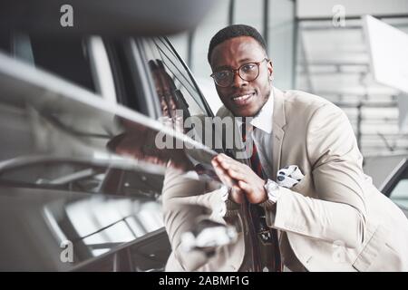 Bel uomo nero in concessionaria è abbracciando la sua nuova vettura e sorridente Foto Stock