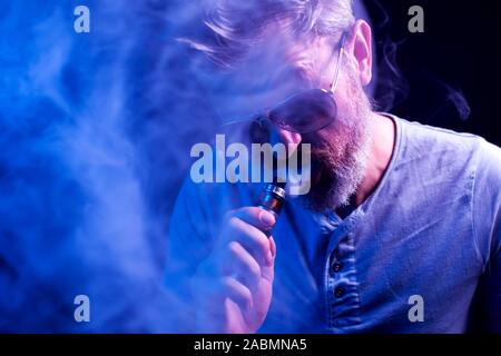 Uomo Barbuto con occhiali da sole fuma vape a sfondo blu. Dipendenza dalle sigarette concept Foto Stock