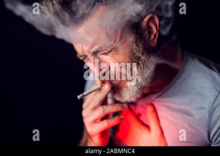 L'uomo fuma sigarette e ha problemi con i polmoni nella parte anteriore del fondo nero. Dipendenza dalle sigarette concept Foto Stock