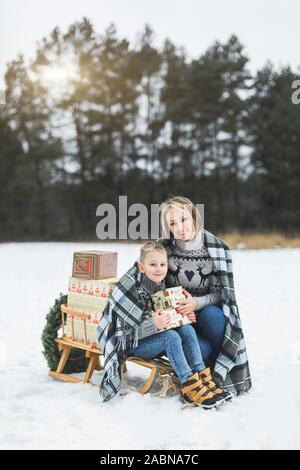 Madre con piccolo figlio in winter park lo slittino. Mamma e Bambino seduto insieme su legno decorato la slitta con i regali di Natale. Passeggiata invernale Foto Stock