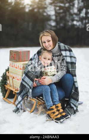 Madre con piccolo figlio in winter park lo slittino. Mamma e Bambino seduto insieme su legno decorato la slitta con i regali di Natale. Passeggiata invernale Foto Stock