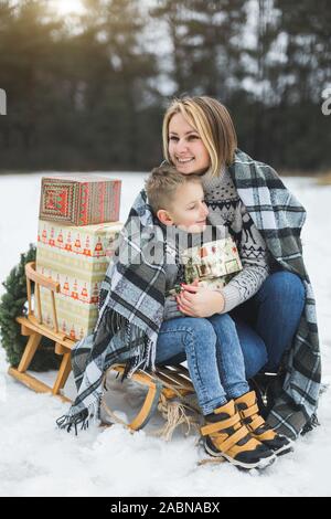 Madre con piccolo figlio in winter park lo slittino. Mamma e Bambino seduto insieme su legno decorato la slitta con i regali di Natale. Passeggiata invernale Foto Stock