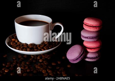 Una tazza di bianco con il nero fragranti caffè sorge su uno sfondo nero con caffè zurnes accanto al francese colorati cookies amaretti Foto Stock