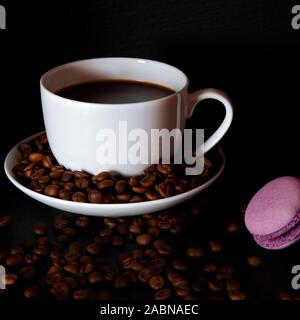 Una tazza di bianco con il nero fragranti caffè sorge su uno sfondo nero con caffè zurnes accanto al francese colorati cookies amaretti Foto Stock