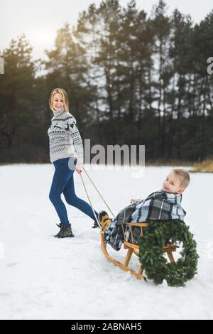 In inverno il divertimento per tutta la famiglia vacanze. Happy mom in felpa lavorata a maglia e jeans porta il suo piccolo figlio su una slitta di legno decorato con ghirlanda di Natale in un inverno Foto Stock