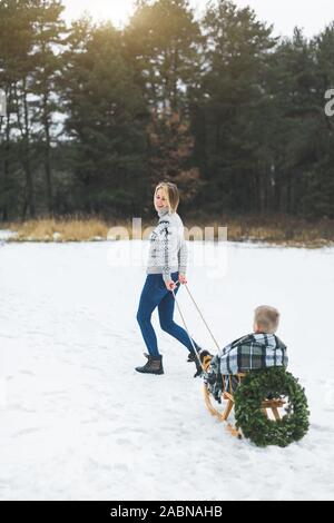 In inverno il divertimento per tutta la famiglia vacanze. Happy mom in felpa lavorata a maglia e jeans porta il suo piccolo figlio su una slitta di legno decorato con ghirlanda di Natale in un inverno Foto Stock