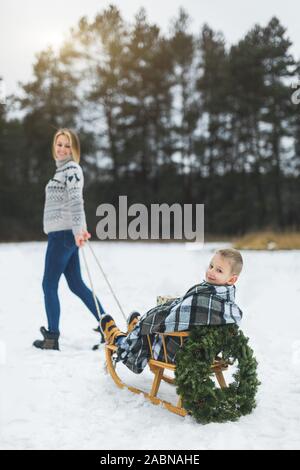 In inverno il divertimento per tutta la famiglia vacanze. Happy mom in felpa lavorata a maglia e jeans porta il suo piccolo figlio su una slitta di legno decorato con ghirlanda di Natale in un inverno Foto Stock