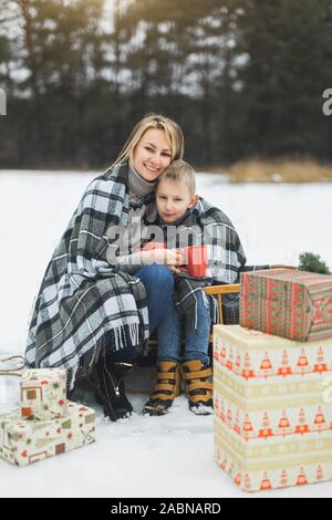 Little Boy e madre scorrevole nella foresta d'inverno. Madre e figlio seduti sulla slitta, di copertura mediante il plaid a scacchi e bere bevande calde Foto Stock