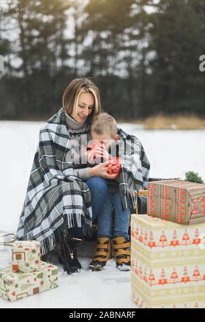 Little Boy e madre scorrevole nella foresta d'inverno. Madre e figlio seduti sulla slitta, di copertura mediante il plaid a scacchi e bere bevande calde Foto Stock