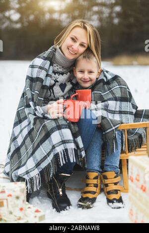 Little Boy e madre scorrevole nella foresta d'inverno. Madre e figlio seduti sulla slitta, di copertura mediante il plaid a scacchi e bere bevande calde Foto Stock