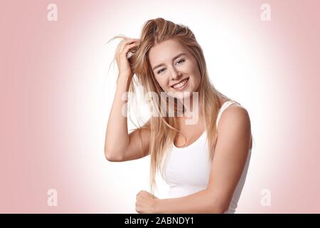 Piuttosto giovane donna bionda con capelli tousled tenendo la mano alla sua testa come sorride alla telecamera oltre a evidenziato sfondo rosa Foto Stock