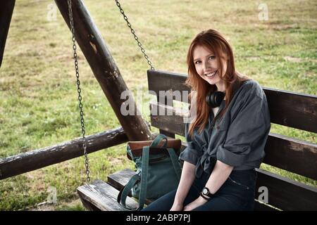 Redhead ragazza in una camicia in denim si siede con uno zaino su una oscillazione di legno da banco e sorrisi nel telaio. Foto Stock