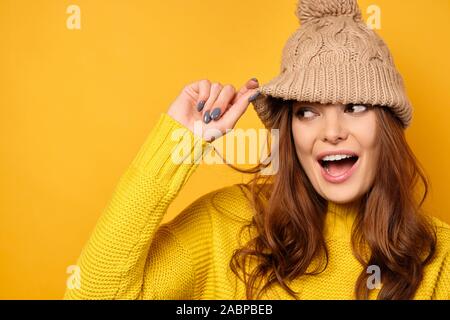 Una bruna in un maglione giallo sorge su uno sfondo giallo e, sollevando un cappello, guarda al lato. Foto Stock