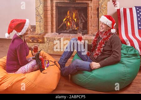 Il marito e la moglie celebrano il nuovo anno. Essi sono qui seduti sul bean bag sedie, bere vino, chiacchierando e sorridendo vicino al camino in Santa's cappelli Foto Stock