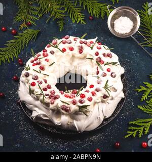 Torta di Natale Pavlova con mirtillo rosso decorato con foglie di rosmarino su sfondo blu scuro accanto a rami di abete. Foto Stock