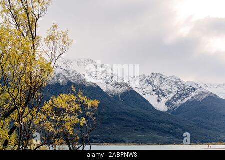 Snow capped montuosa incorniciato dalla giallo fogliame di un albero in un lago con un cielo nuvoloso Foto Stock