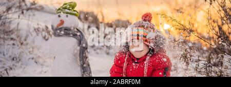 Carino boy in rosso abbigliamento invernale costruisce un pupazzo di neve. Divertimento invernale Outdoor Concept banner, formato lungo Foto Stock