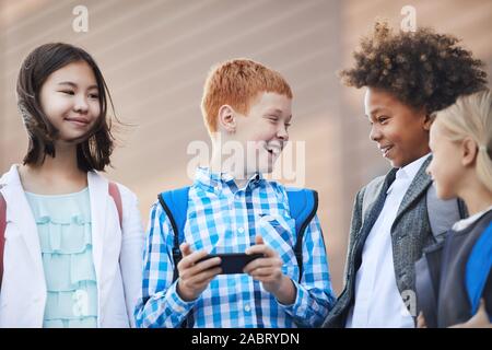 Dai capelli rossi boy in piedi con il telefono cellulare e ridere mentre parla ai suoi compagni di classe all'aperto Foto Stock