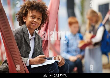 Ritratto di ragazzo africano con capelli ricci guardando la fotocamera durante la scrittura in notebook all'aperto nel parco giochi Foto Stock