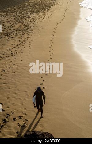 Un surfista passeggiate su una spiaggia di sabbia al tramonto, lasciando dietro di sé le impronte sulla sabbia dorata Foto Stock