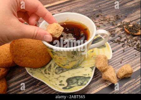 La mano della ragazza mette un pezzo di zucchero di canna in una tazza di tè nero, fiocchi d'avena cookies, foglie di tè e zucchero di canna su uno sfondo di legno. Close up Foto Stock