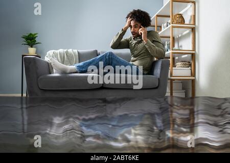 Allagato il pavimento dalla perdita di acqua nella parte anteriore del uomo seduto sul divano chiamando per idraulico Foto Stock