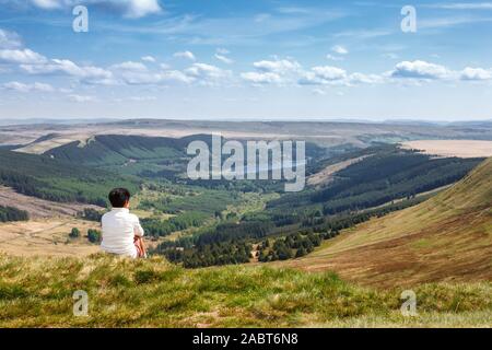 Indian donna seduta su di una collina nella campagna gallese guardando a valle e verso l'orizzonte. Pen Y ventola, Brecon Beacons, South Wales, Regno Unito Foto Stock