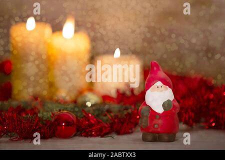 Il calore del concetto di Natale: un Babbo Natale statuetta in primo piano in un ambiente rustico con tre candele accese in bokeh effetto, oro e r Foto Stock