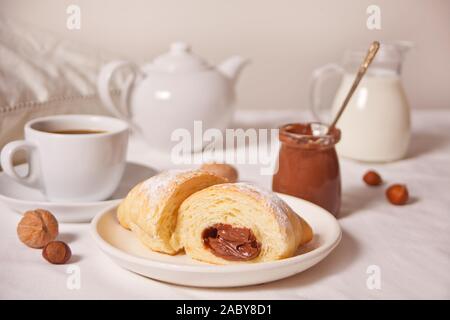 Cornetti freschi bun con cioccolato sulla piastra, una tazza di caffè, un vasetto di latte nelle vicinanze sullo sfondo bianco. Foto Stock