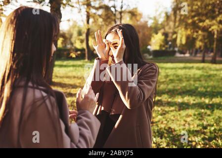 Due brunette gemelle in piedi di fronte ad ogni altro divertendosi e facendo dei cerchi con le dita intorno agli occhi, indossando cappotto casual in autunno sunny Foto Stock