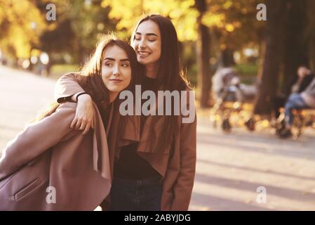 Ritratto di giovane sorridente brunette due gemelle abbracciando e divertirsi in cappotto casual in piedi vicino all'altra in autunno soleggiato parco alley su sfocata Foto Stock