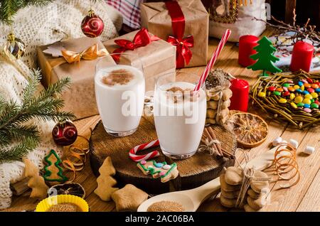 Natale e Anno Nuovo zabaione bevande in bicchieri di vetro su un supporto di legno sul tavolo con i regali di Natale i cookie, candele, dolciumi e albero di Natale bra Foto Stock