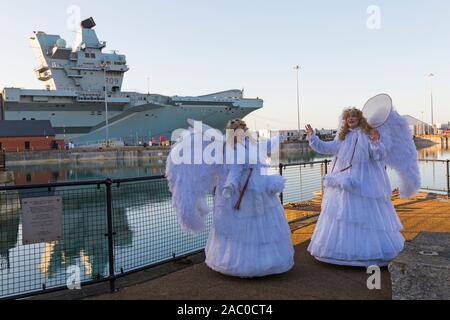 Portsmouth, Hampshire, Regno Unito. 29 novembre 2019. Le folle affollano il festival vittoriano di Natale a Portsmouth Historic Dockyard per l'intrattenimento, i personaggi vestiti in olden giorni e il mercato di Natale, il primo giorno dell'evento di tre giorni. Angeli con la portaerei HMS Prince of Wales dietro. Credit: Carolyn Jenkins/Alamy Live News Foto Stock