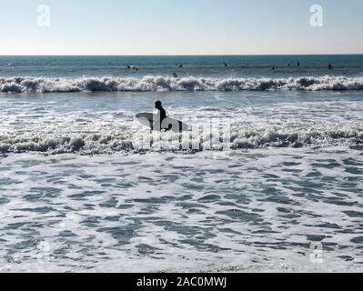 In inverno il surf in spiaggia Selsey nel febbraio 2019 con un surfista camminare nelle onde tenendo la sua tavola da surf Foto Stock