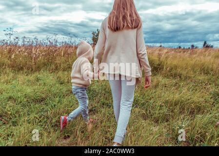 Donna madre cammina campo, trattiene la mano del ragazzino figlio, città d'autunno, passeggiata nel parco, erba di sfondo e un caldo abbigliamento informale, beige cardigan con cappuccio Foto Stock